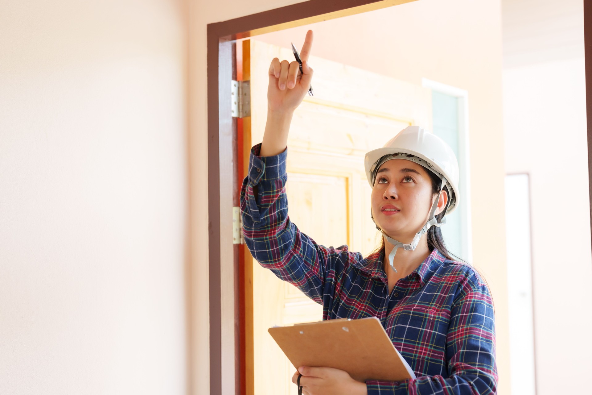 Professional Asian inspector in plaid shirt, white safety helmet, assessing house quality, clipboard and orange tape measure in hand, examining doorway. Inspectors inspect the completed house